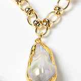 Close-up detail of a luminous baroque pearl pendant framed in a sculptural gold-plated bezel and suspended from a polished circular-link chain with metallic accents and refined artisanal detailing.