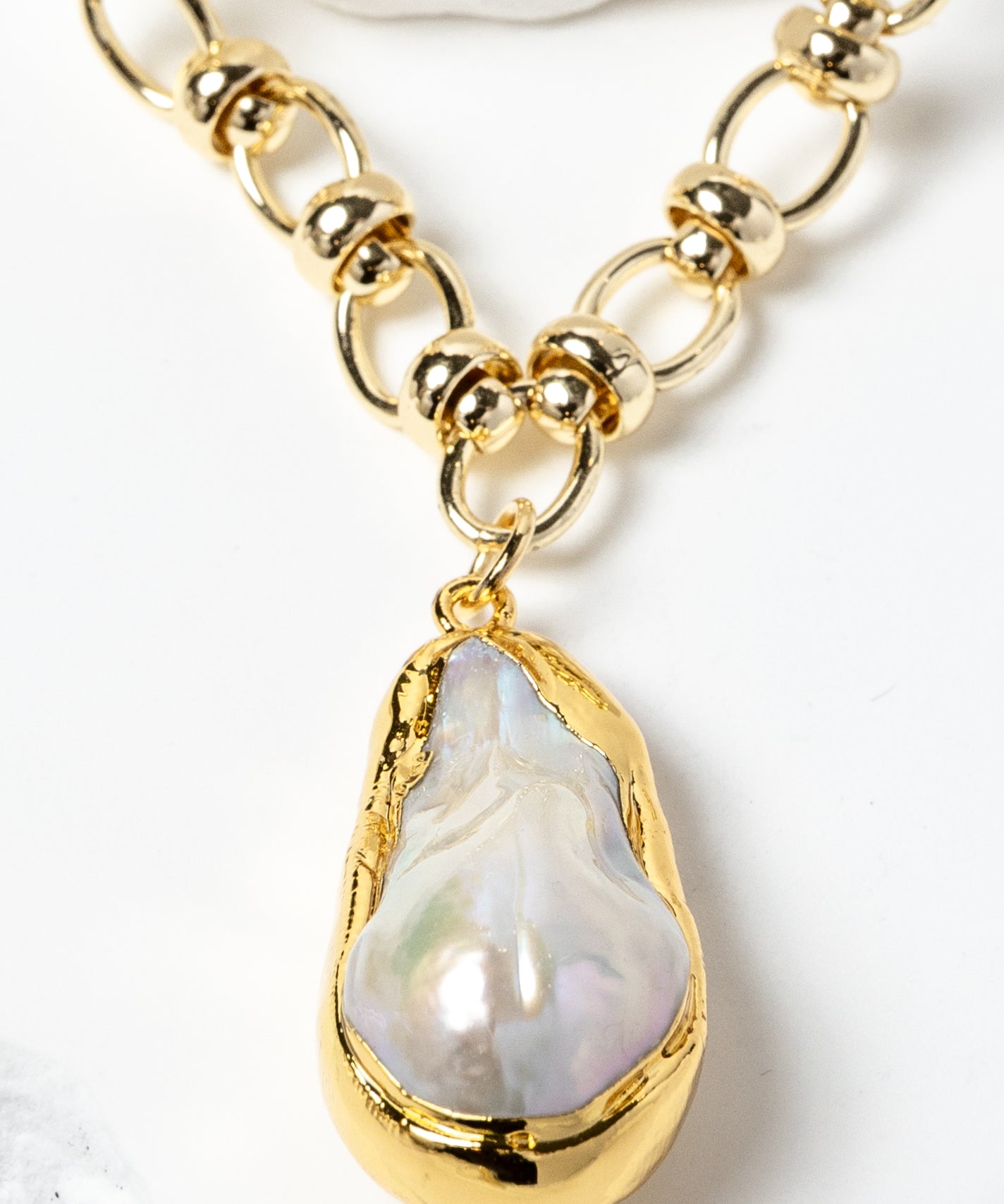 Close-up detail of a luminous baroque pearl pendant framed in a sculptural gold-plated bezel and suspended from a polished circular-link chain with metallic accents and refined artisanal detailing.