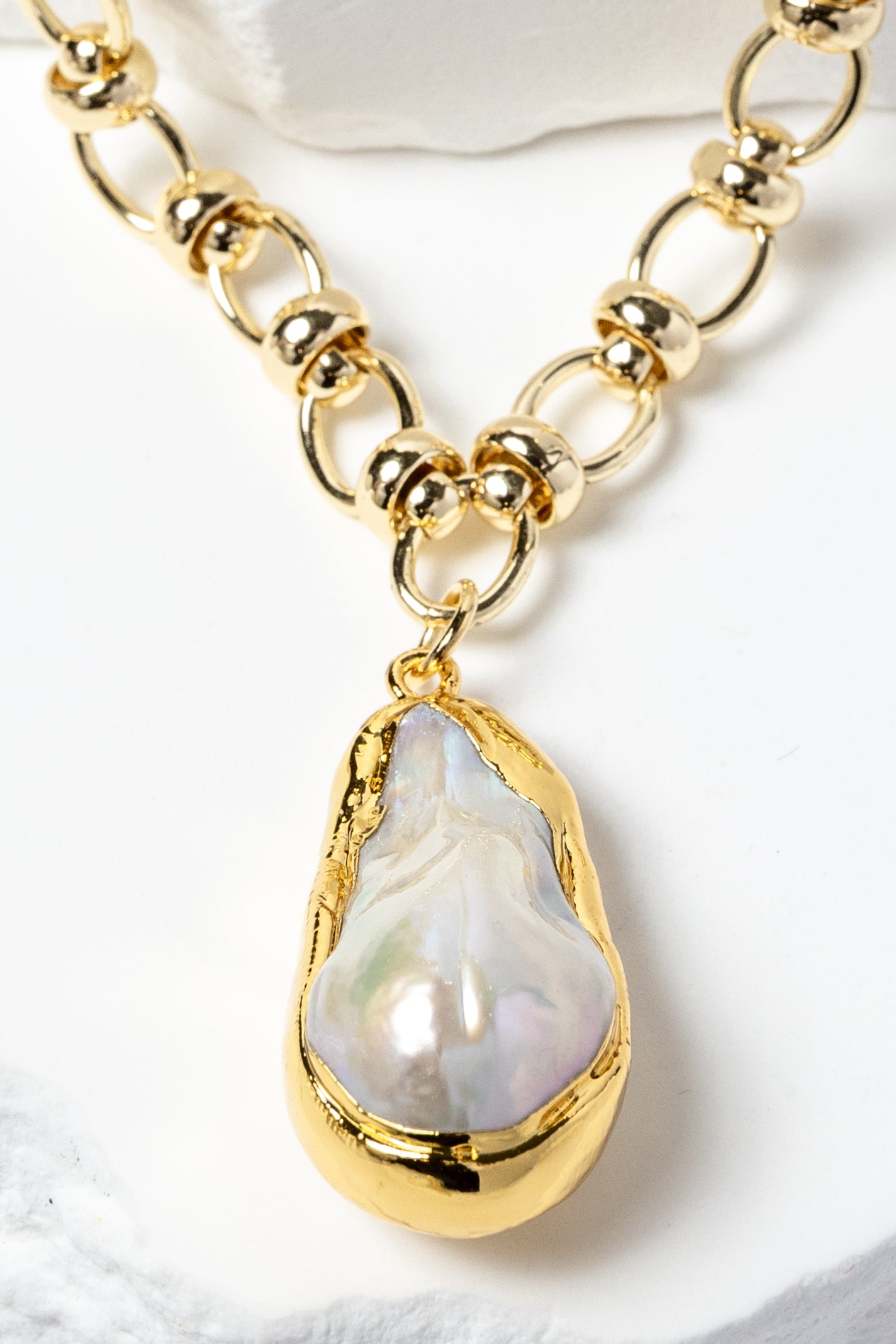 Close-up detail of a luminous baroque pearl pendant framed in a sculptural gold-plated bezel and suspended from a polished circular-link chain with metallic accents and refined artisanal detailing.