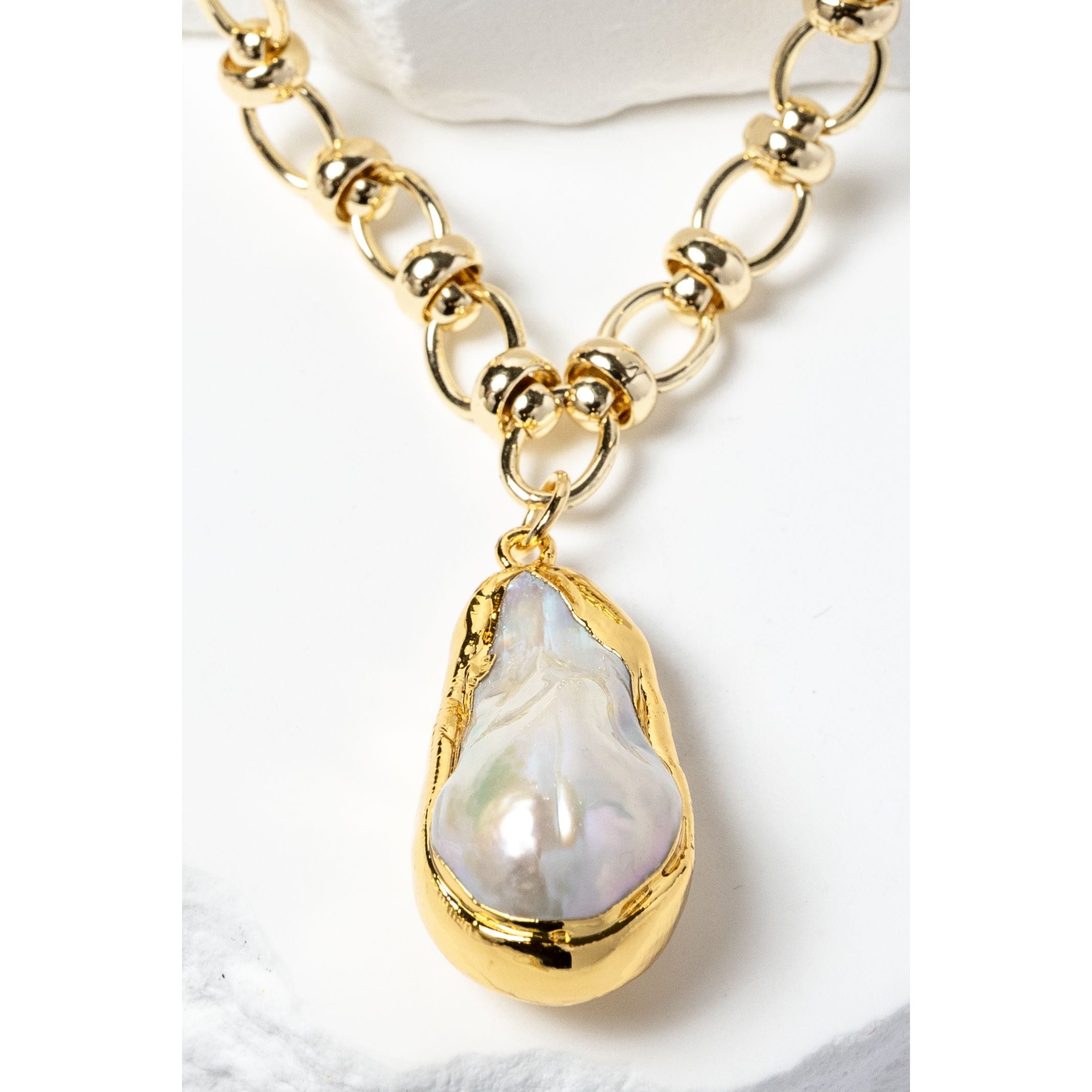Close-up detail of a luminous baroque pearl pendant framed in a sculptural gold-plated bezel and suspended from a polished circular-link chain with metallic accents and refined artisanal detailing.