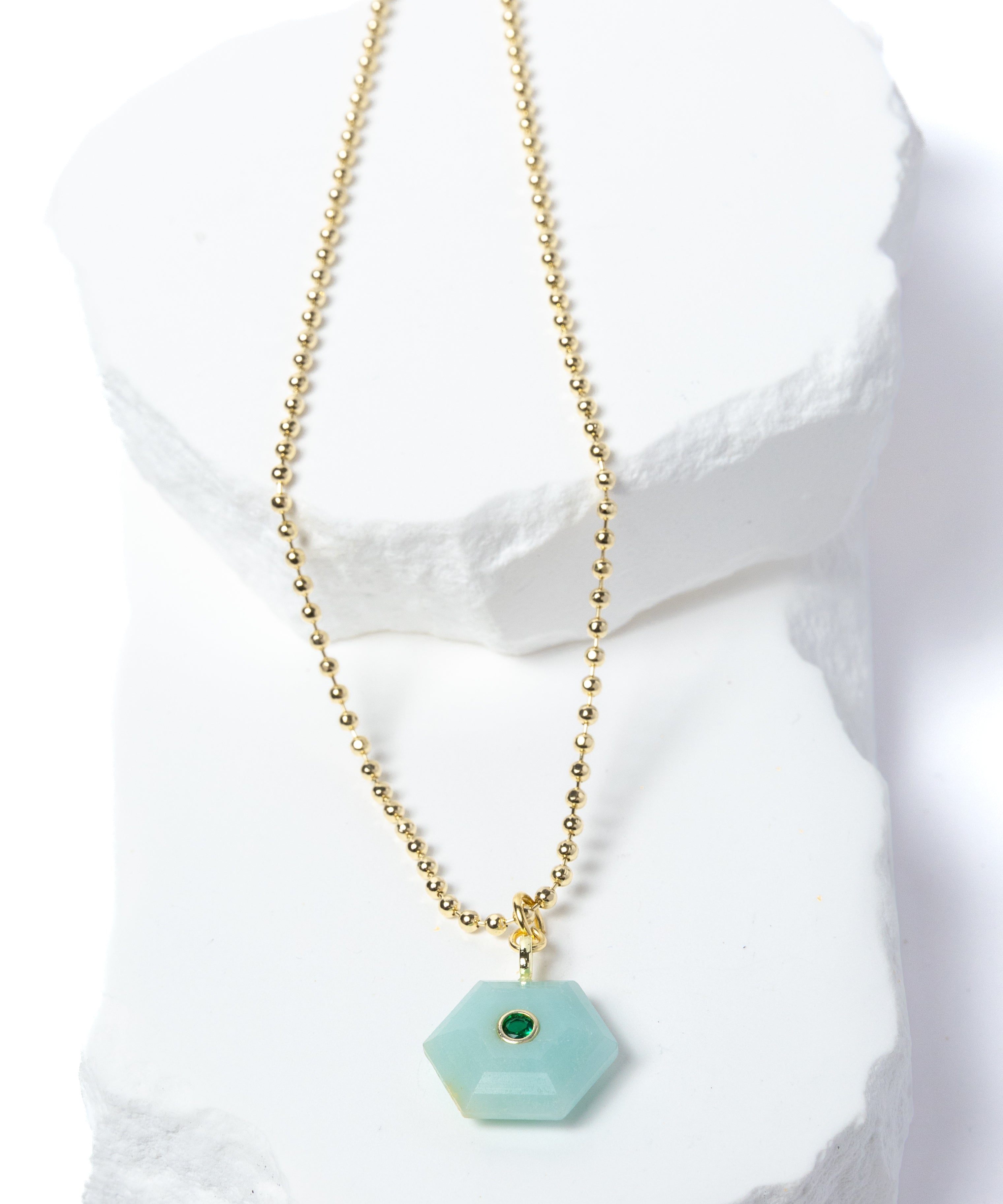 Natural Amazonite Gem Charm Necklace featuring a faceted soft aqua gemstone pendant with a green cubic zirconia accent suspended from a gold-plated beaded chain displayed on a white sculptural stone backdrop.