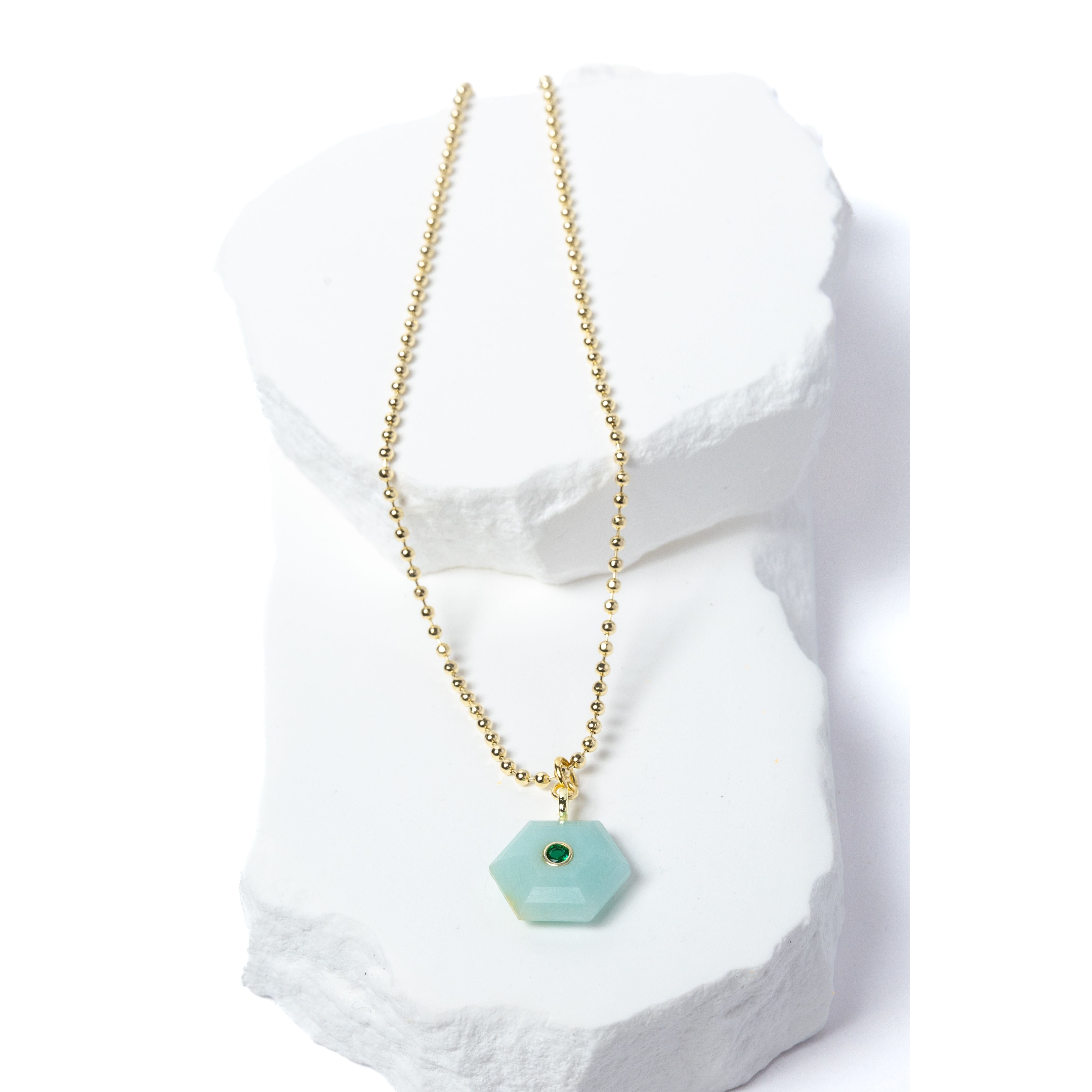 Natural Amazonite Gem Charm Necklace featuring a faceted soft aqua gemstone pendant with a green cubic zirconia accent suspended from a gold-plated beaded chain displayed on a white sculptural stone backdrop.