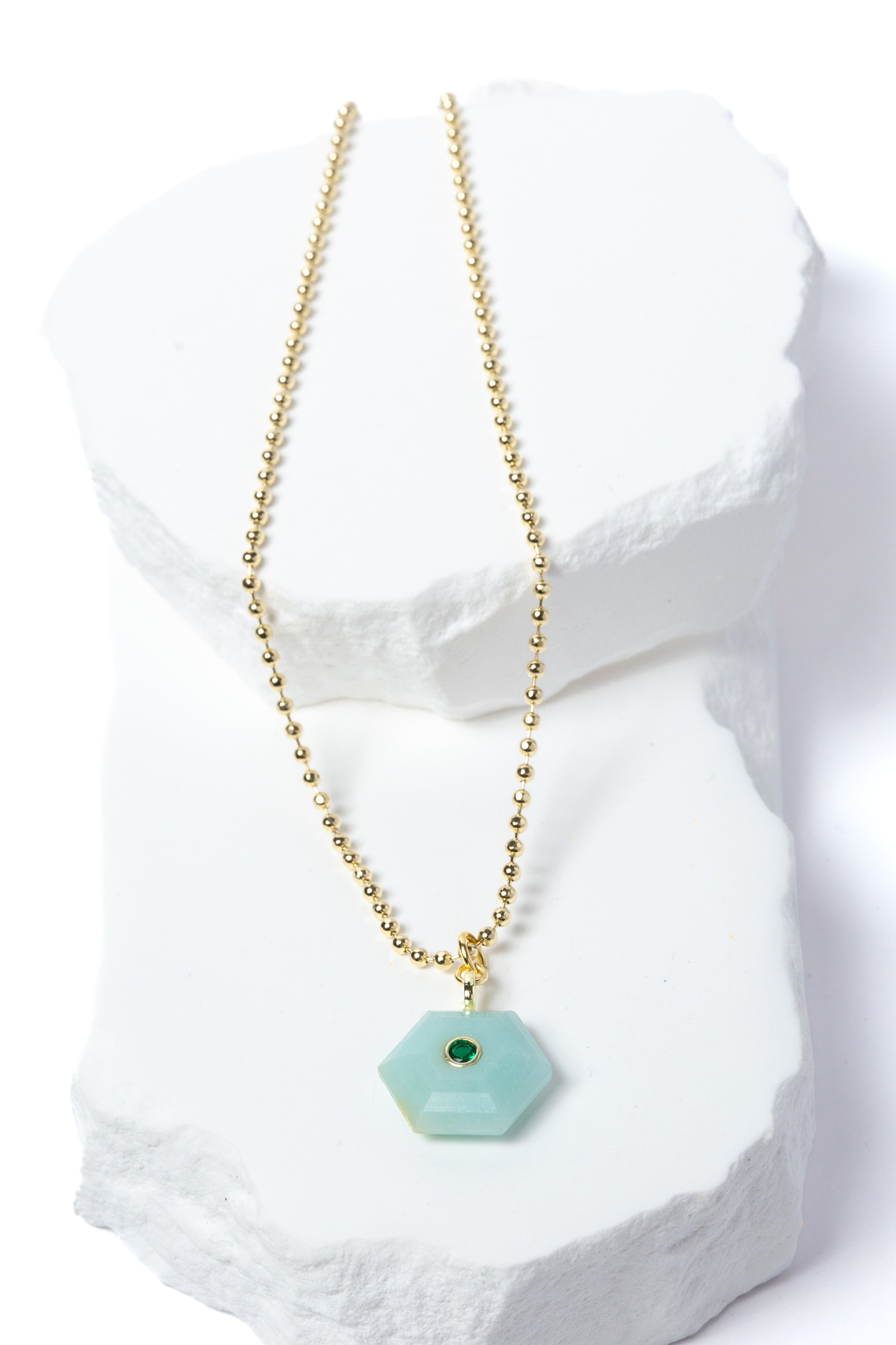 Natural Amazonite Gem Charm Necklace featuring a faceted soft aqua gemstone pendant with a green cubic zirconia accent suspended from a gold-plated beaded chain displayed on a white sculptural stone backdrop.