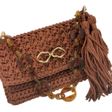 Close-up of the Polly Clutch in a rich cinnamon-brown tone featuring dimensional handcrafted crochet texture, translucent amber-inspired resin chain strap, polished gold-tone front accent, and elongated tassel detail with sculptural movement.