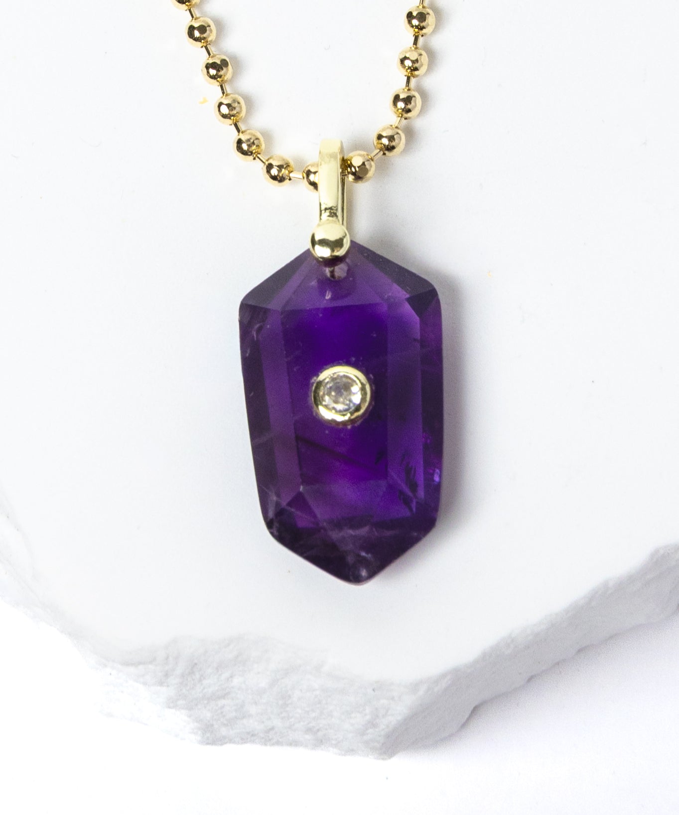 Close-up of a natural Amethyst pendant showcasing rich violet-purple tones, faceted geometric shape, cubic zirconia detail, and gold-plated setting.