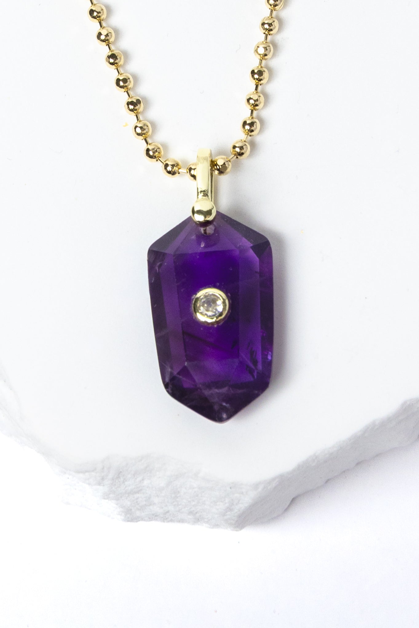 Close-up of a natural Amethyst pendant showcasing rich violet-purple tones, faceted geometric shape, cubic zirconia detail, and gold-plated setting.
