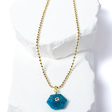 Natural Apatite Gem Charm Necklace featuring a vibrant ocean-blue faceted gemstone pendant with a cubic zirconia accent suspended from a gold-plated beaded chain displayed on a white sculptural stone backdrop.