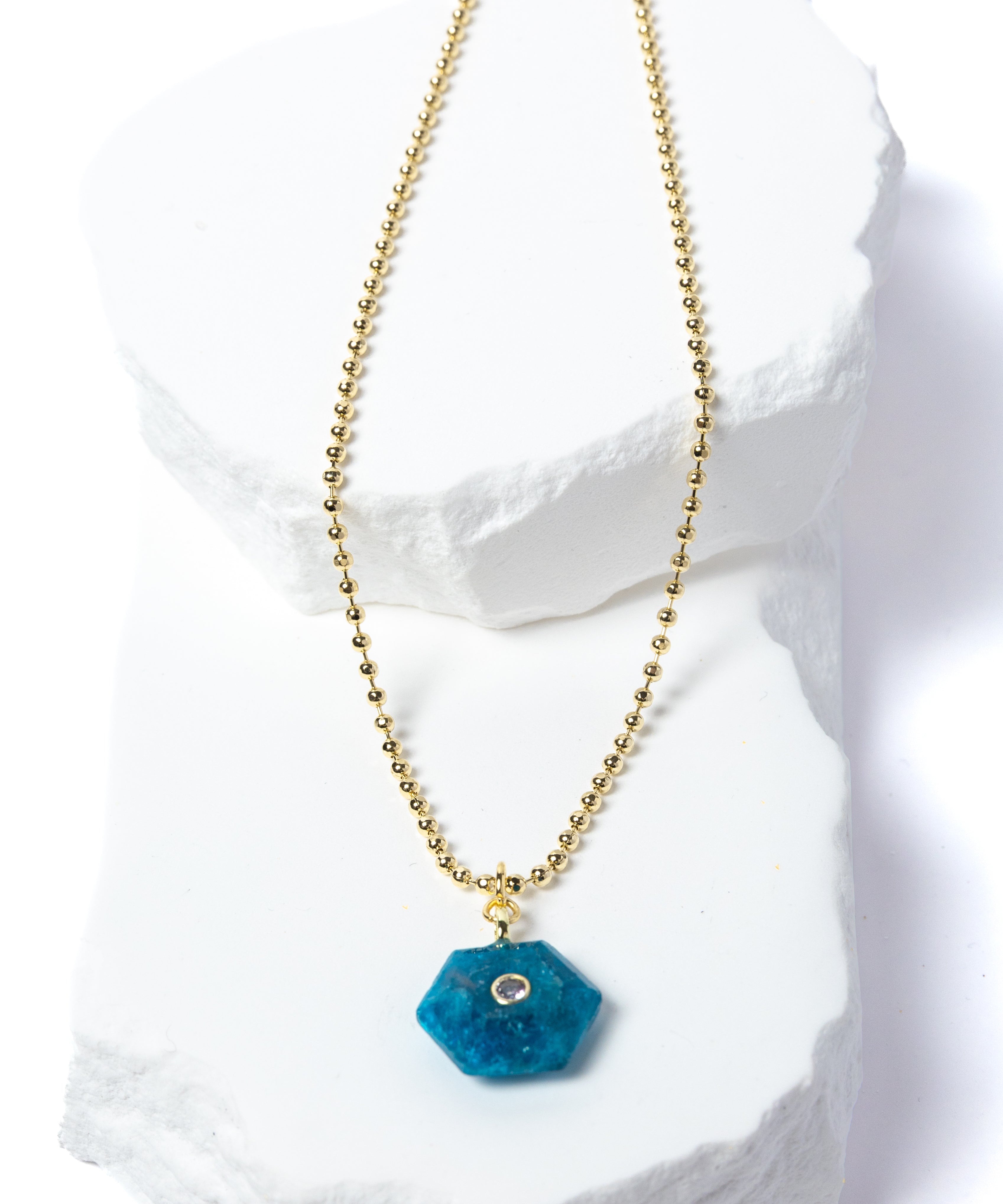 Natural Apatite Gem Charm Necklace featuring a vibrant ocean-blue faceted gemstone pendant with a cubic zirconia accent suspended from a gold-plated beaded chain displayed on a white sculptural stone backdrop.