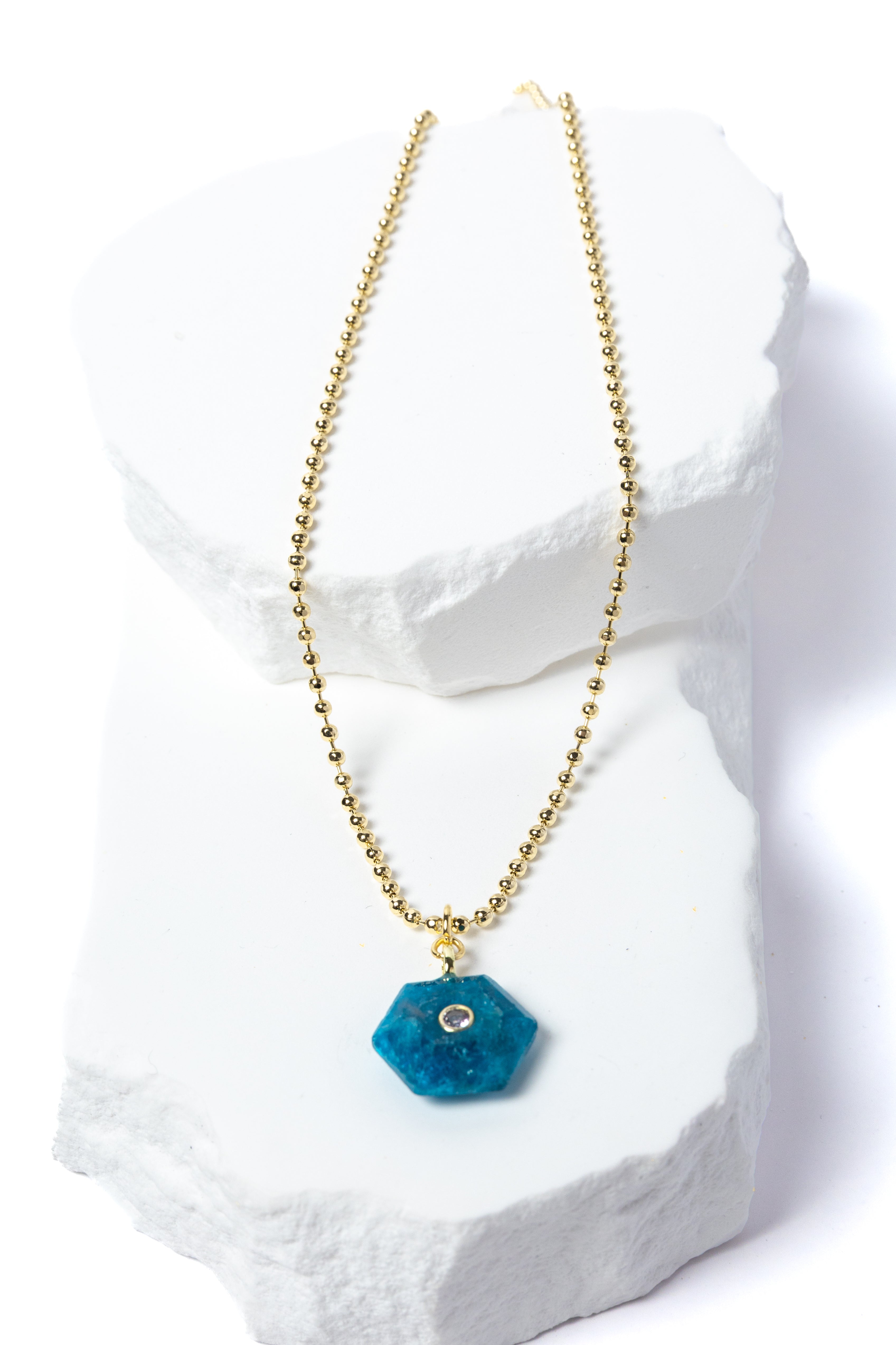 Natural Apatite Gem Charm Necklace featuring a vibrant ocean-blue faceted gemstone pendant with a cubic zirconia accent suspended from a gold-plated beaded chain displayed on a white sculptural stone backdrop.