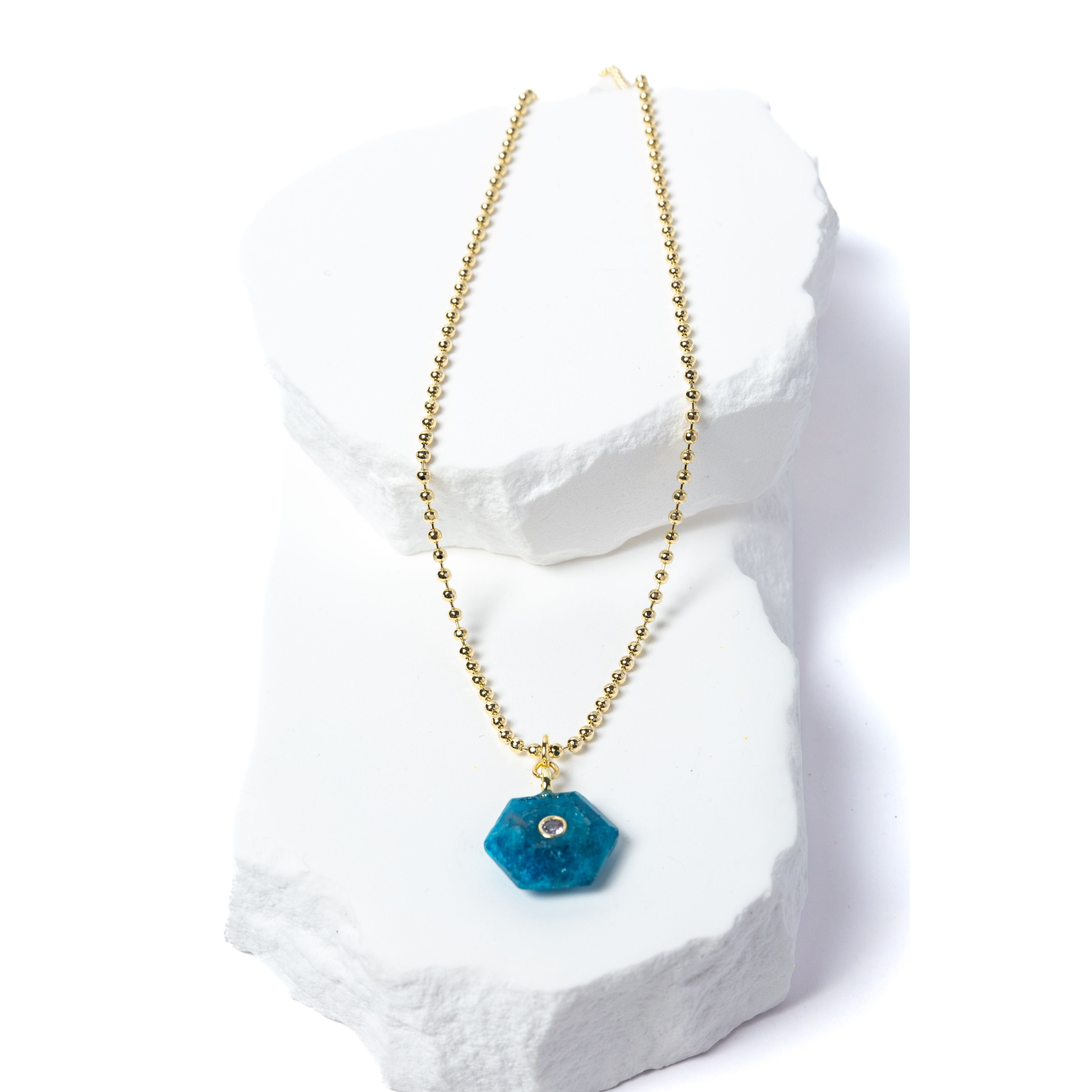 Natural Apatite Gem Charm Necklace featuring a vibrant ocean-blue faceted gemstone pendant with a cubic zirconia accent suspended from a gold-plated beaded chain displayed on a white sculptural stone backdrop.