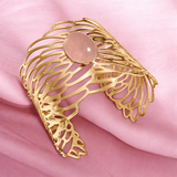 Golden Symphony Cuff with a polished oval rose quartz cabochon in soft blush pink, surrounded by flowing, sculptural golden openwork in a wide statement silhouette.