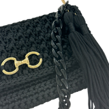 Angled view of the Polly Clutch Ebony Edition showcasing the textured black crochet, resin chain strap, decorative gold-tone front accent, and elegant tassel.