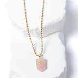 Natural Rose Quartz Gem Charm Necklace featuring a faceted blush-pink gemstone pendant with a cubic zirconia accent suspended from a gold-plated beaded chain displayed on a white sculptural stone backdrop.