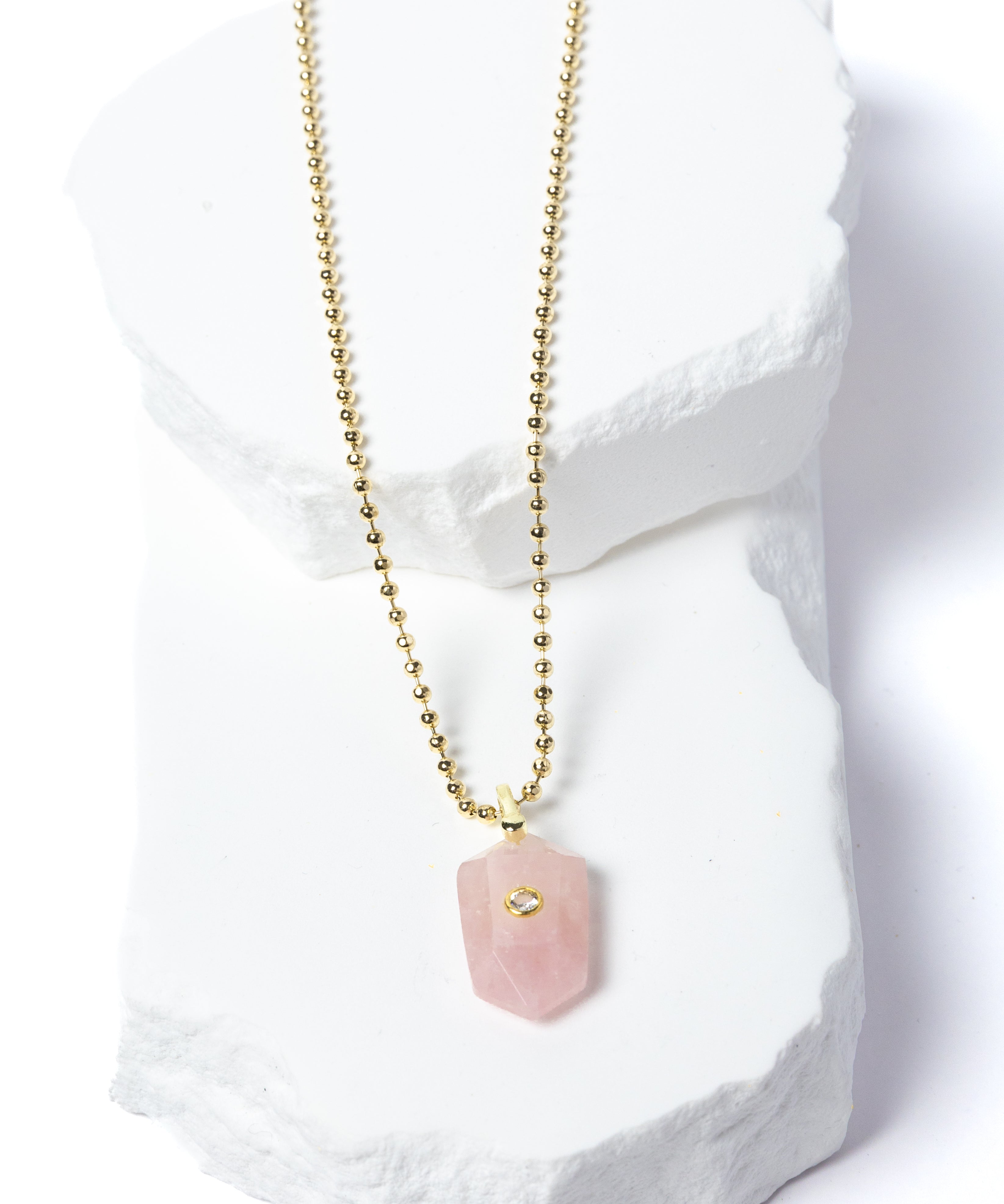 Natural Rose Quartz Gem Charm Necklace featuring a faceted blush-pink gemstone pendant with a cubic zirconia accent suspended from a gold-plated beaded chain displayed on a white sculptural stone backdrop.