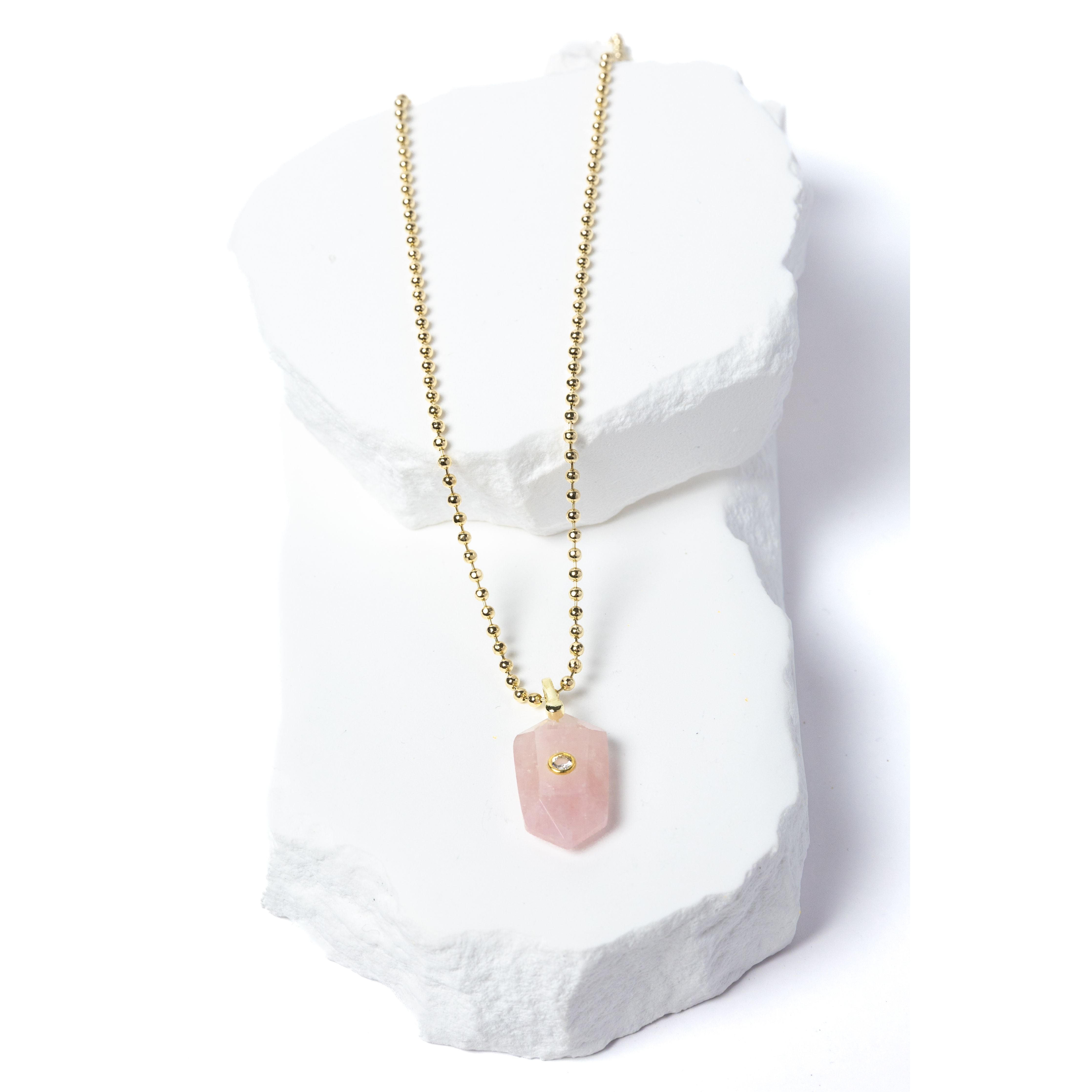 Natural Rose Quartz Gem Charm Necklace featuring a faceted blush-pink gemstone pendant with a cubic zirconia accent suspended from a gold-plated beaded chain displayed on a white sculptural stone backdrop.