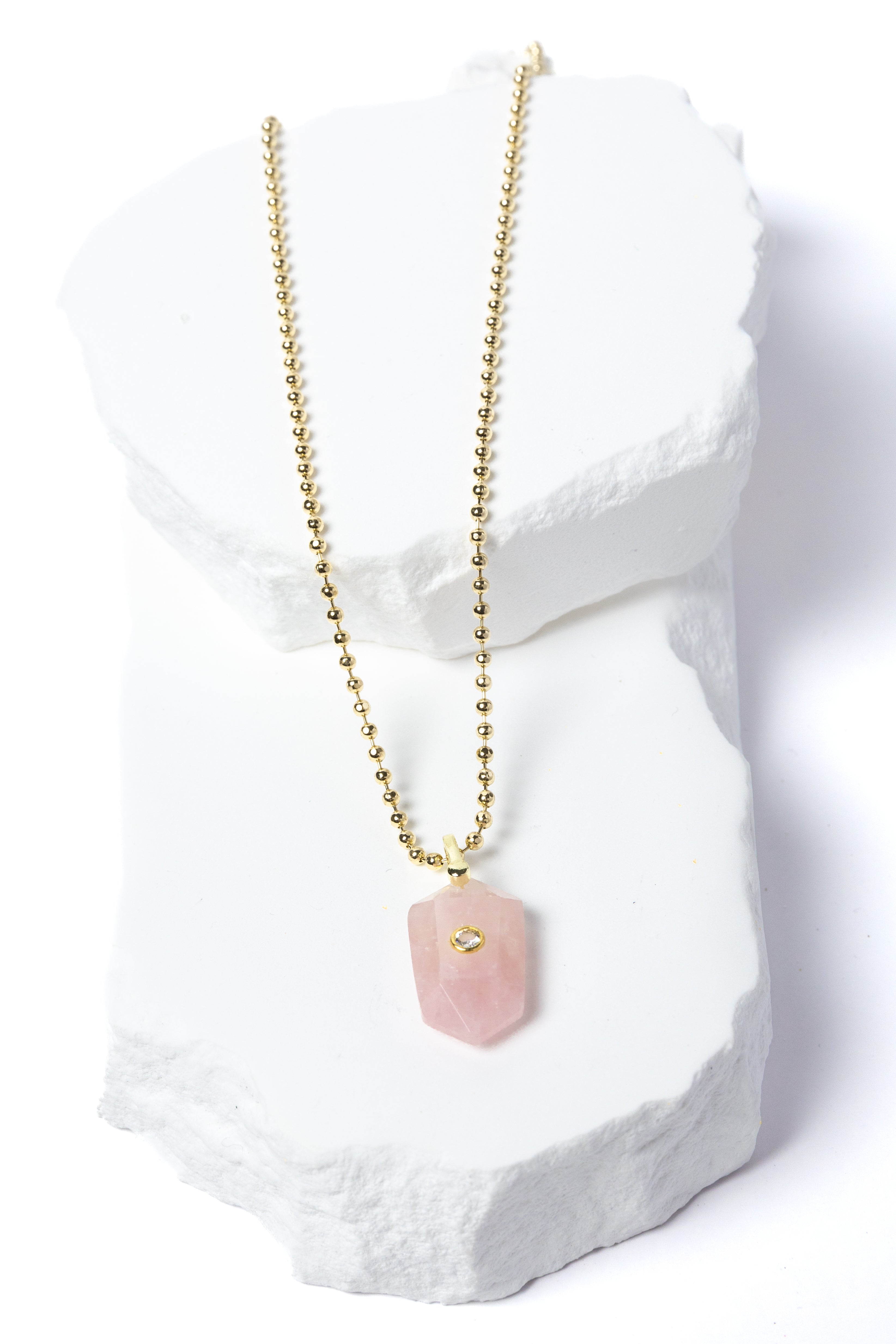 Natural Rose Quartz Gem Charm Necklace featuring a faceted blush-pink gemstone pendant with a cubic zirconia accent suspended from a gold-plated beaded chain displayed on a white sculptural stone backdrop.