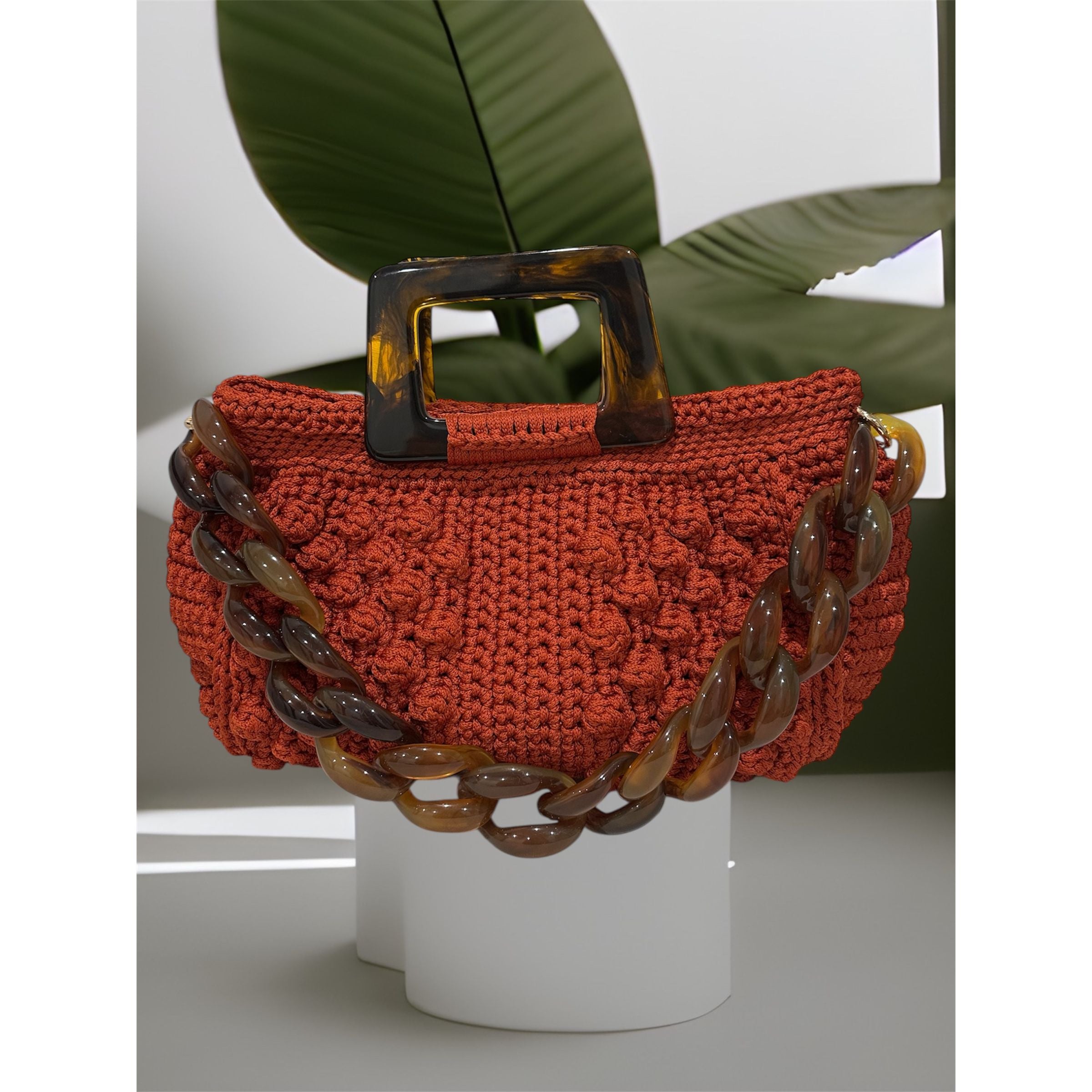 Hand-crocheted Paloma handbag in a burnt terracotta tone with dimensional textured stitching, featuring a marbled amber resin top handle and a coordinating chain strap.