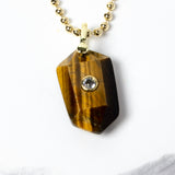 Close-up of a natural Tiger Eye pendant showcasing golden-brown banding, faceted geometric shape, cubic zirconia detail, and gold-plated setting.