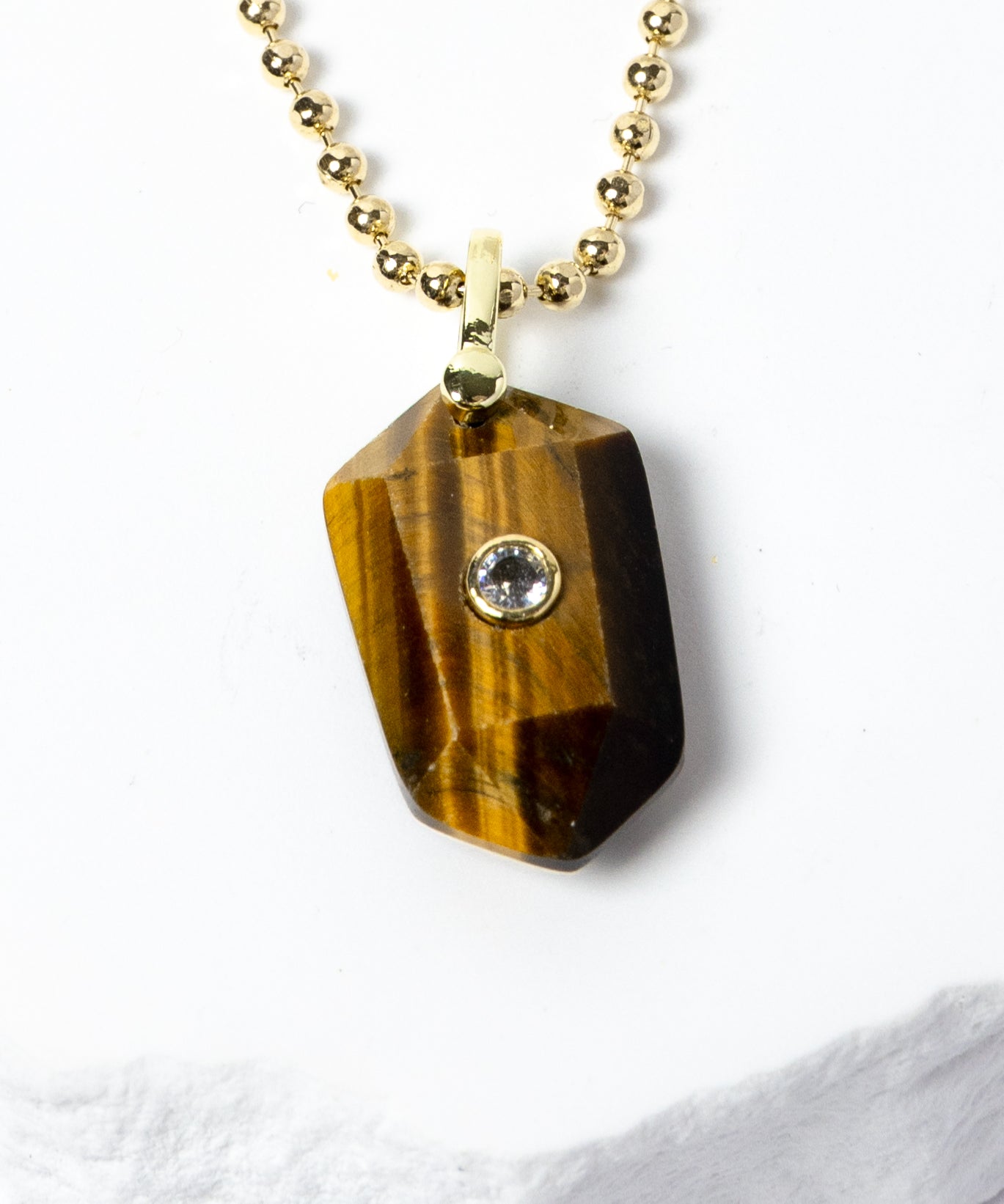 Close-up of a natural Tiger Eye pendant showcasing golden-brown banding, faceted geometric shape, cubic zirconia detail, and gold-plated setting.