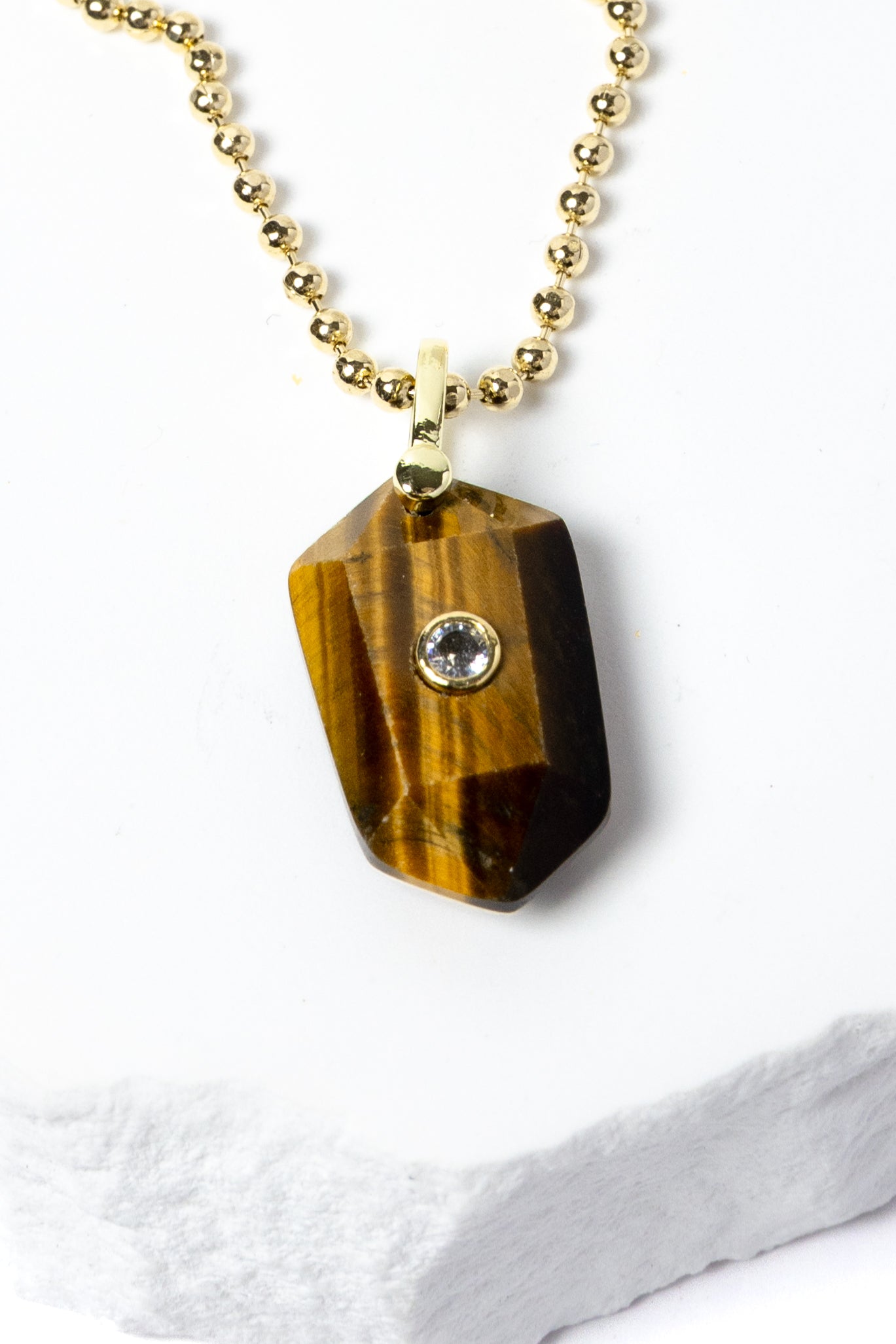 Close-up of a natural Tiger Eye pendant showcasing golden-brown banding, faceted geometric shape, cubic zirconia detail, and gold-plated setting.